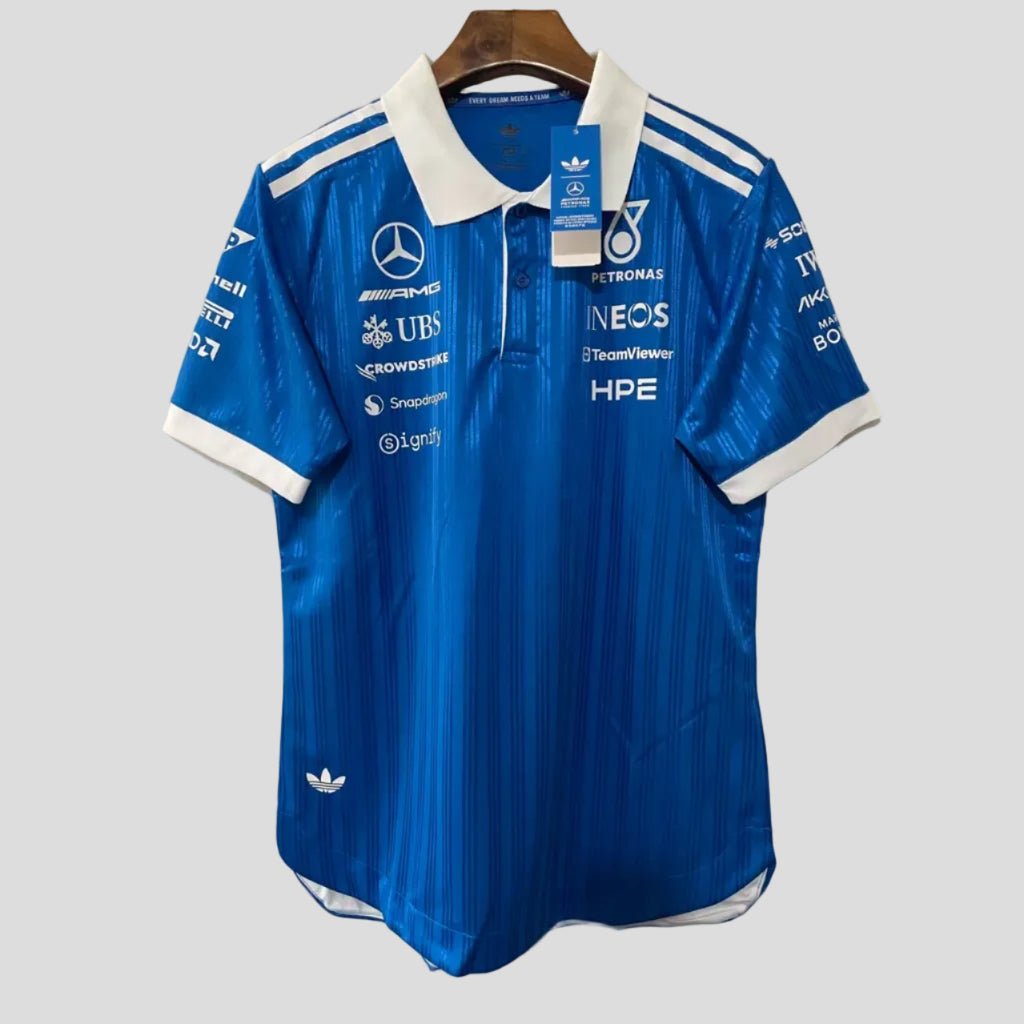 A blue Mercedes Petronas Formula 1 racing polo shirt by Adidas, featuring white sponsor logos and branding, hanging on a wooden hanger against a white brick wall.