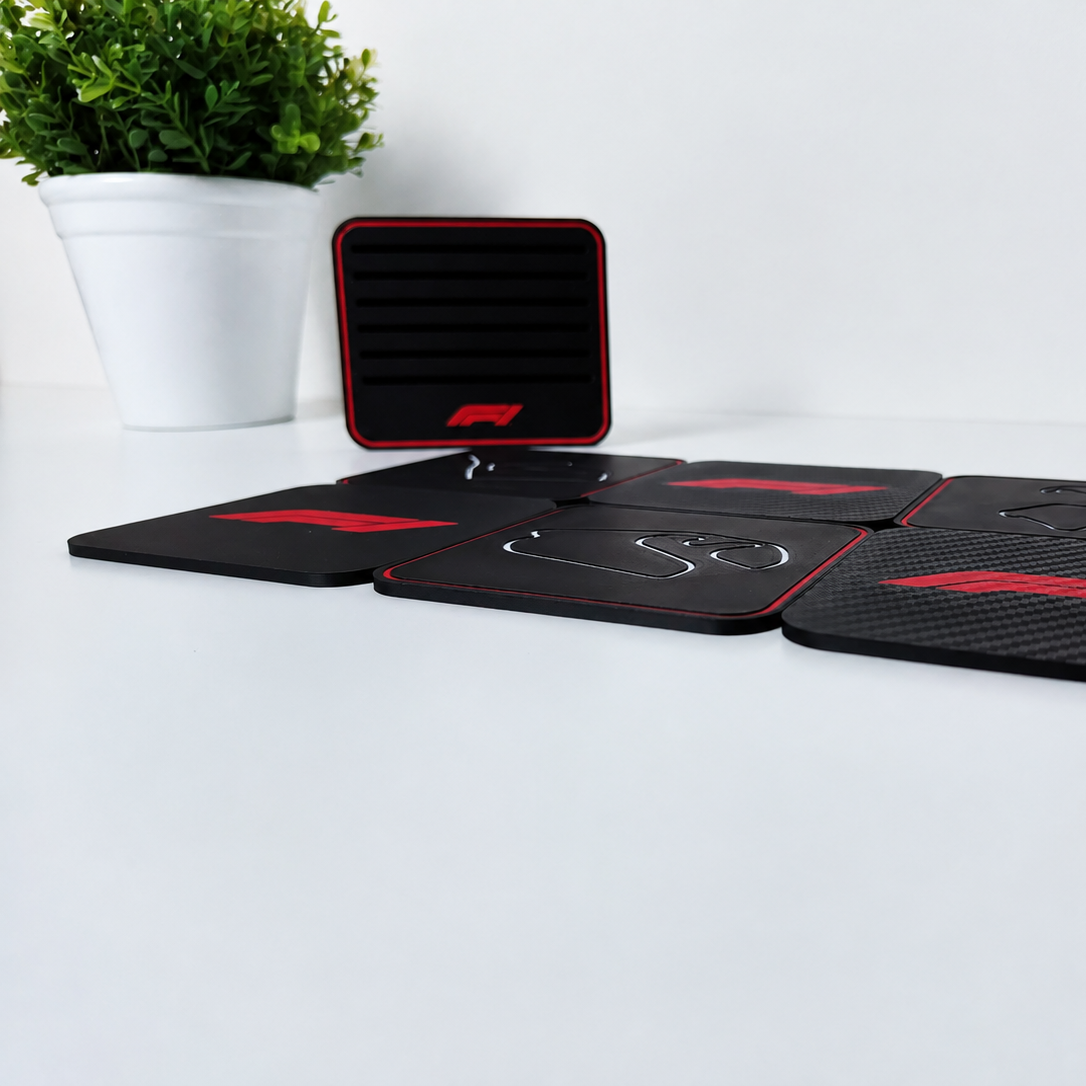 A set of black rubber coasters with red trim and white F1-style logos, displayed on a white surface with a small potted plant in the background.