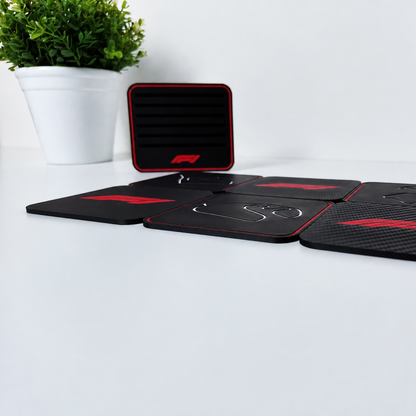 A set of black rubber coasters with red trim and white F1-style logos, displayed on a white surface with a small potted plant in the background.