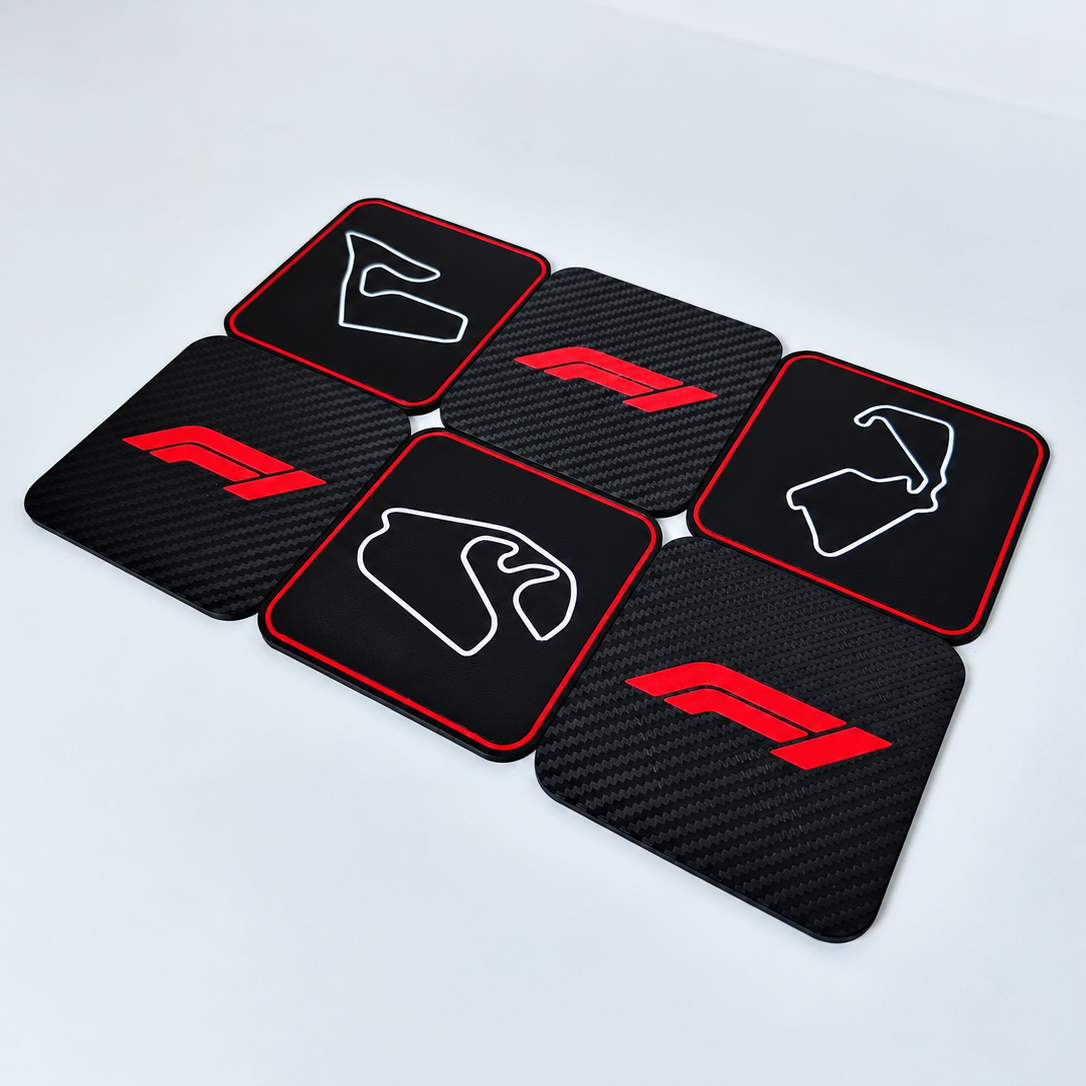 Six black square coasters with rounded corners arranged in a loose cluster on a light gray surface. The coasters feature a carbon fiber texture, red borders, and red and white F1-style logos, including a stylized horse head and circuit map outlines.