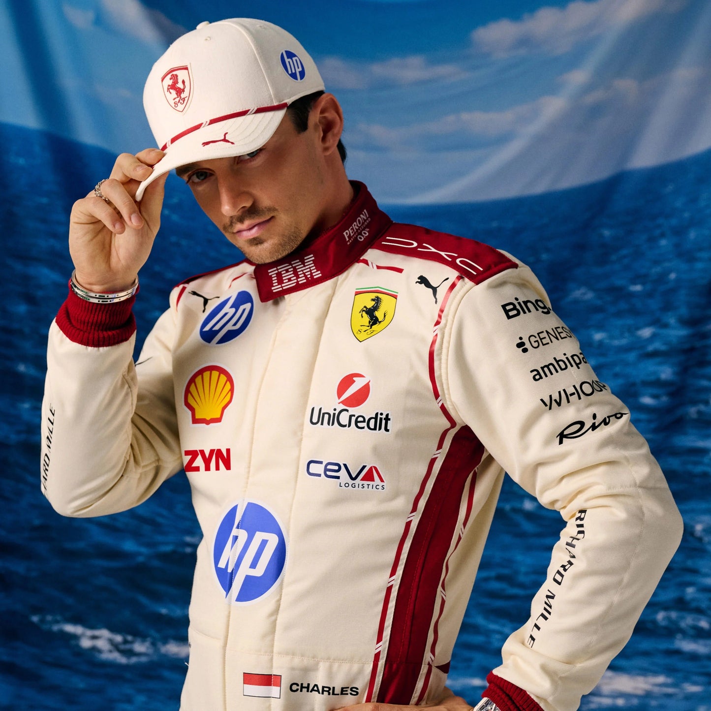 Charles Leclerc in a white and red Ferrari racing suit with sponsor logos, wearing a matching white Ferrari cap and posing against a blue ocean backdrop.